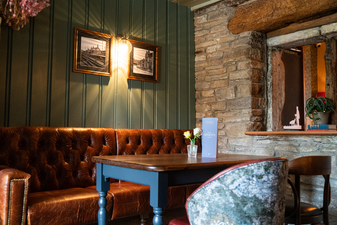 The Snug at The Farmers Boy. Exposed wall to the back right, panelled wall painted in a heritage green with rustic leather banquet seating and a table